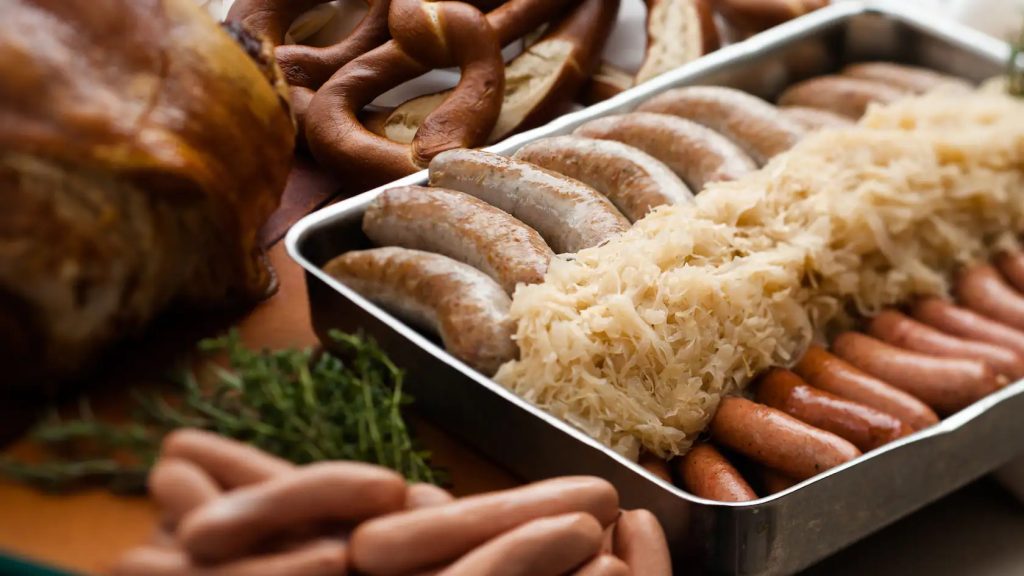 Pan filled with sauerkraut and 2 types of German sausages sitting on a table next to meats and pretzels - Biergarten Restaurant